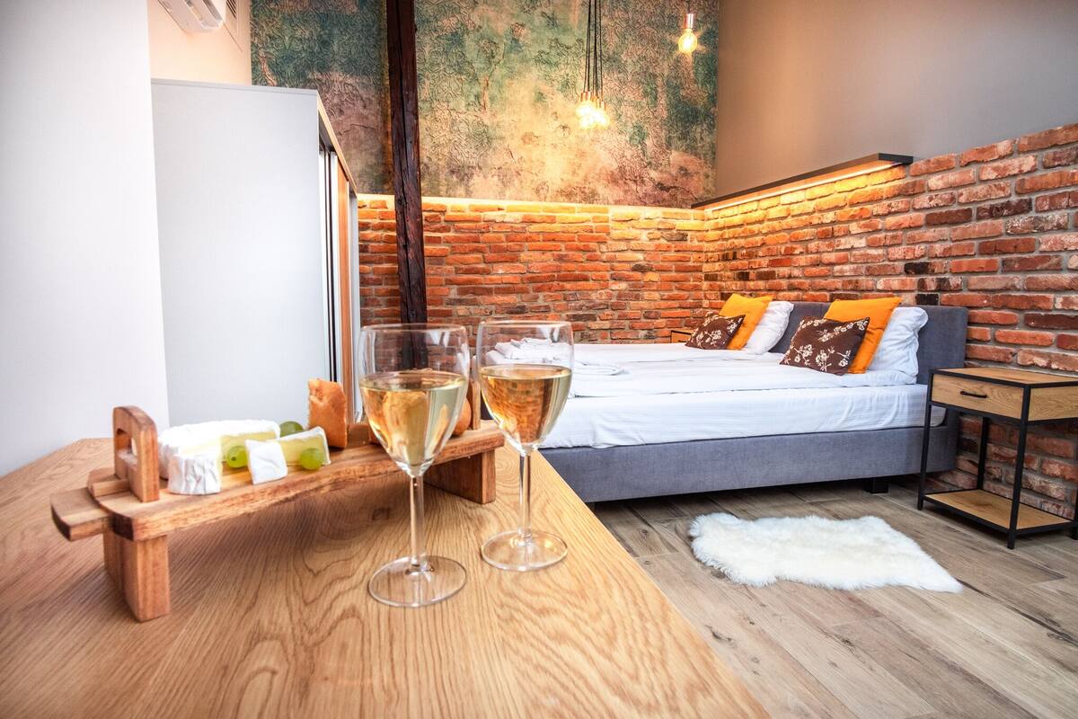 A modern room with a brick wall, a bed with orange pillows, a wooden table with glasses and cheese, and a white fur rug.