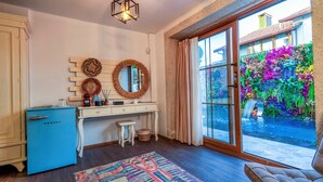 Um quarto com uma escrivaninha de madeira, uma cómoda azul, um espelho e vista para um jardim colorido através de uma porta de vidro.