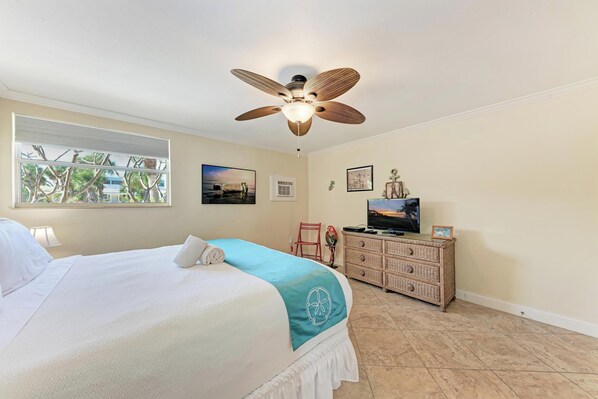 A bedroom with a bed, a ceiling fan, a dresser, a TV, and a window with a view of palm trees. - Catchin' Rays-150 yards to the Beach-By Island Real Estate Vacations (Holmes Beach)