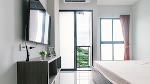 Una camera da letto moderna con un letto, una TV fissata al muro, una piccola scrivania con una pianta e una porta scorrevole in vetro che dà su un balcone.