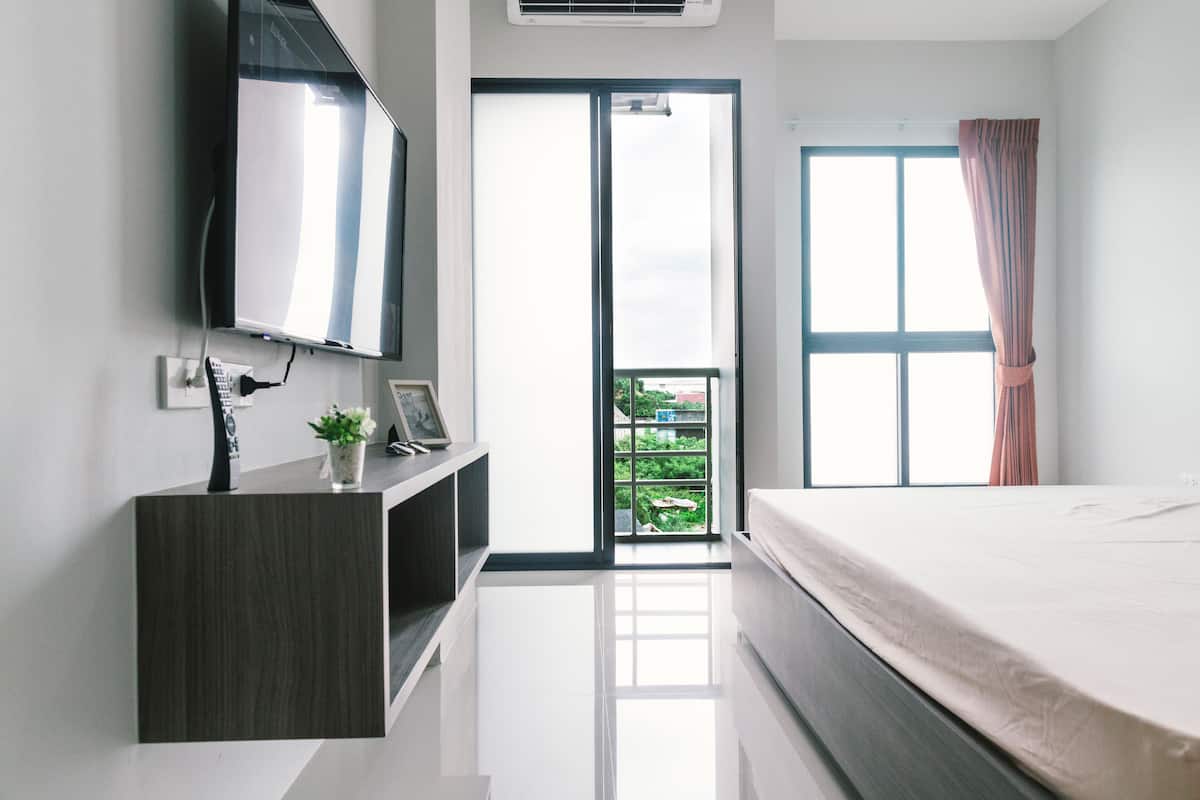 Ein modernes Schlafzimmer mit Bett, einem an der Wand befestigten Fernseher, einem kleinen Schreibtisch mit Pflanze und einer Glastür, die auf einen Balkon führt.