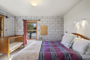 Een slaapkamer met een houten wieg, een bed met gestreept beddengoed, een raam met uitzicht en een prikbord aan de muur.