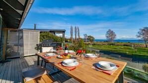 Un espace extérieur aménagé pour les repas, avec une table pour quatre personnes, un barbecue et une vue panoramique sur l’océan et les collines.