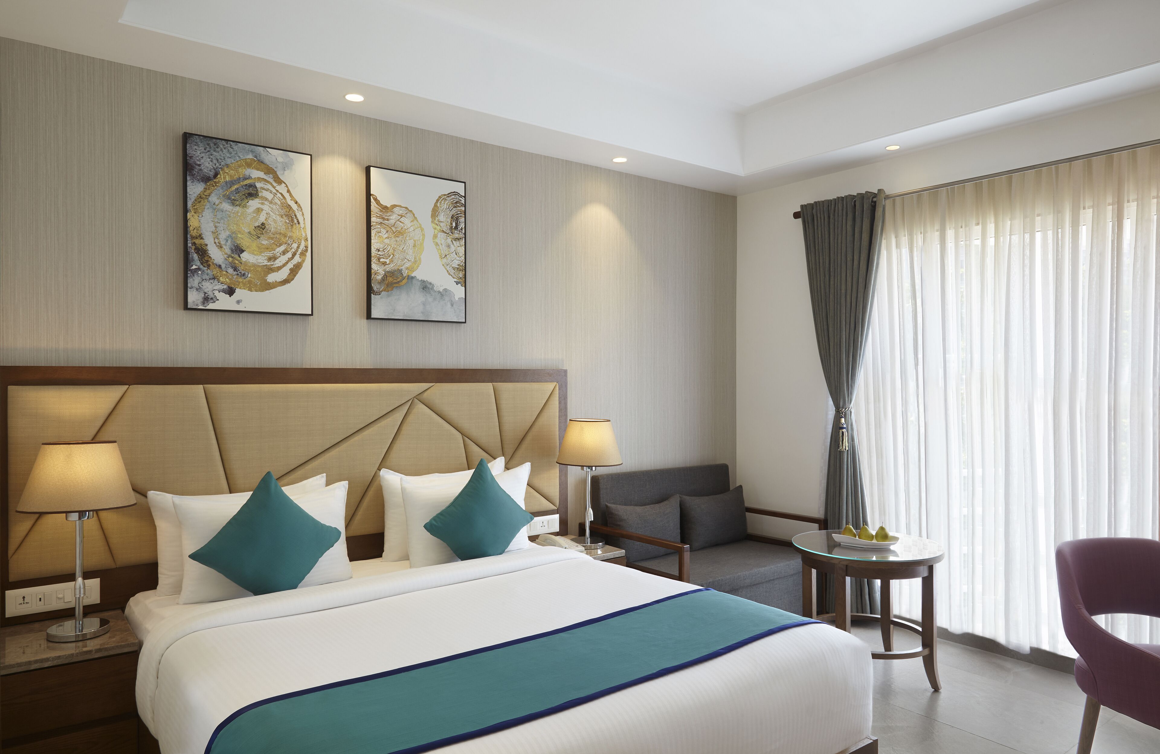 Una camera d'hotel con un letto grande, due lampade ai comodini, un tavolino con una ciotola di frutta, una sedia e due quadri astratti alle pareti.