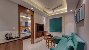 A modern living room with a teal sofa, a small wooden table with a tray of items, a television mounted on the wall, and a ceiling fan. - Click Hotel Aurangabad (Chhatrapati Sambhajinagar)