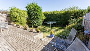 Une terrasse en bois avec une table et des chaises, entourée de plantes en pot et d’une haie.