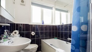 A bathroom with a white sink, toilet, and bathtub, black tiled walls, and a window with blinds. - Woodstock (Poole)
