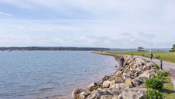Une promenade en bord de mer, avec une bordure rocheuse, une zone herbeuse et une vue sur la mer, où l’on aperçoit des bateaux au loin.