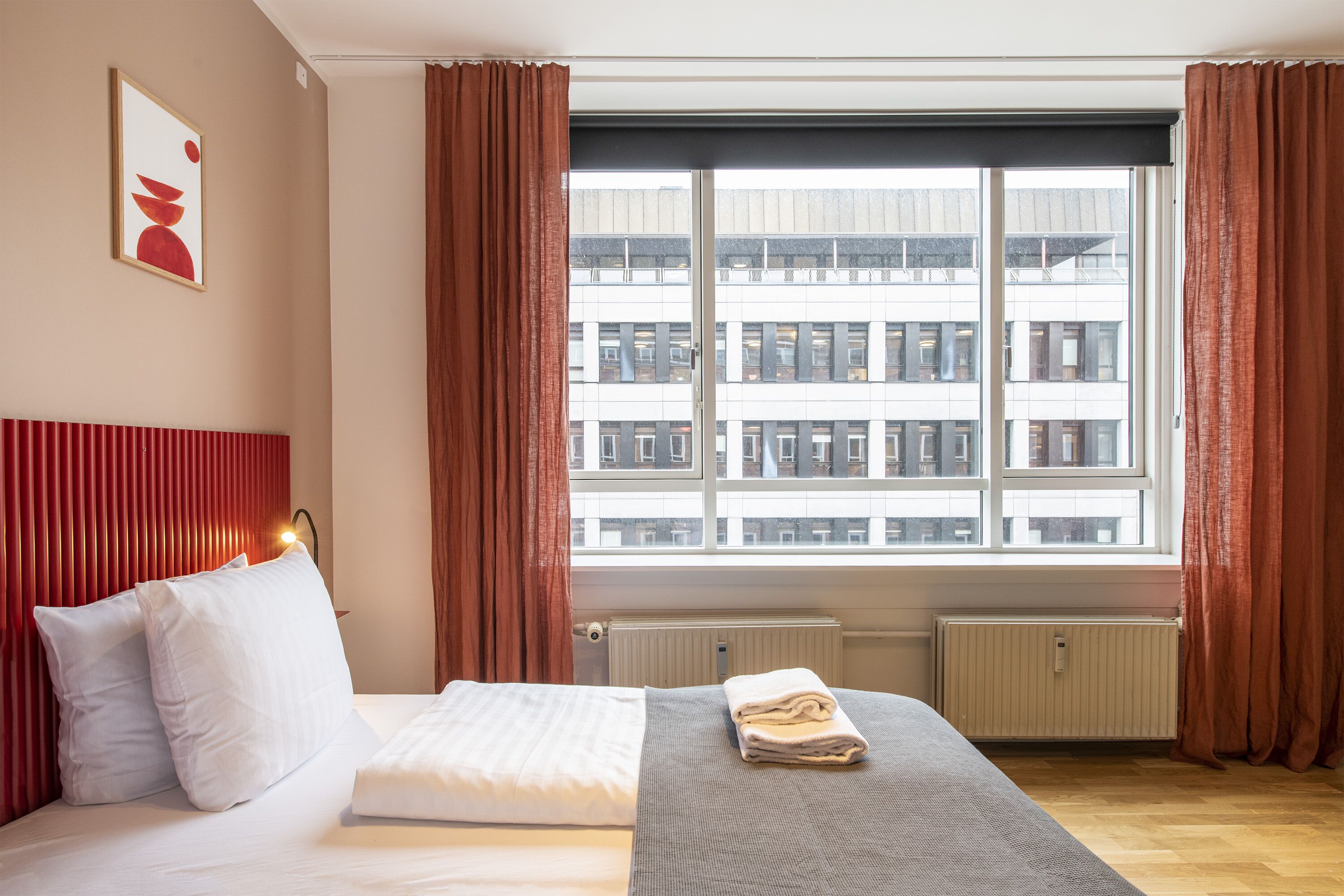 Quarto de hotel com uma cama, uma janela com vista para edifícios e um radiador.