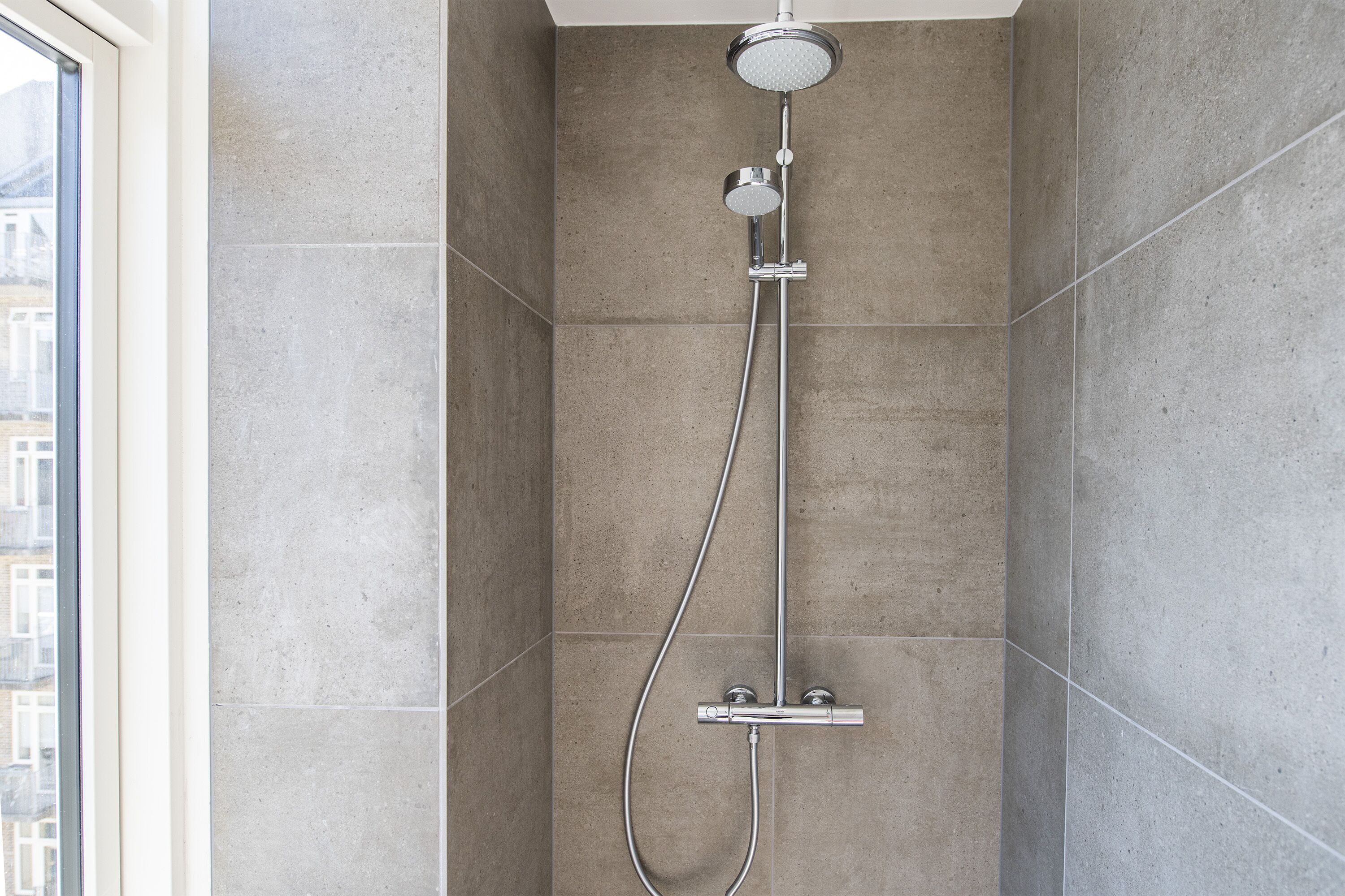 Une douche moderne équipée d’un pommeau de douche à main et d’un pommeau de douche fixe au plafond, le tout installé sur un mur recouvert de grandes carreaux de couleur claire.