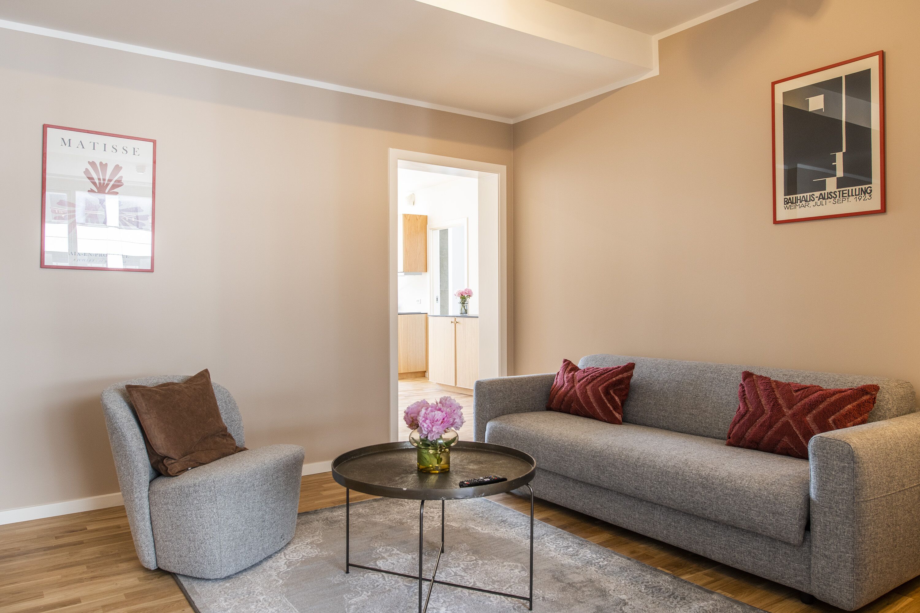 A living room with a grey sofa, a grey armchair, a small round coffee table with a vase of flowers, and a framed artwork on the wall.
