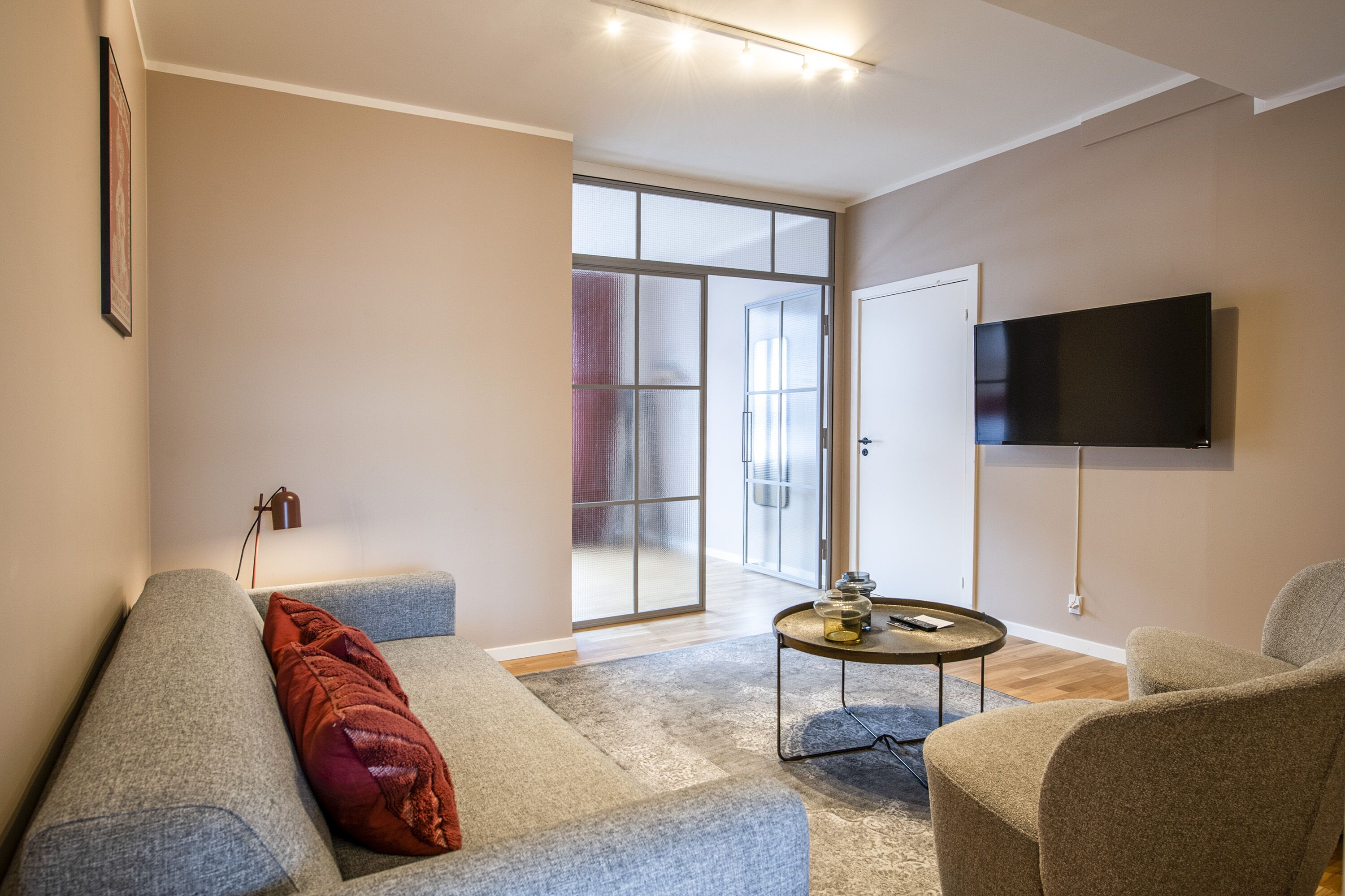 A modern living room with a grey sofa, a red throw pillow, a coffee table, and a flat-screen TV mounted on the wall.