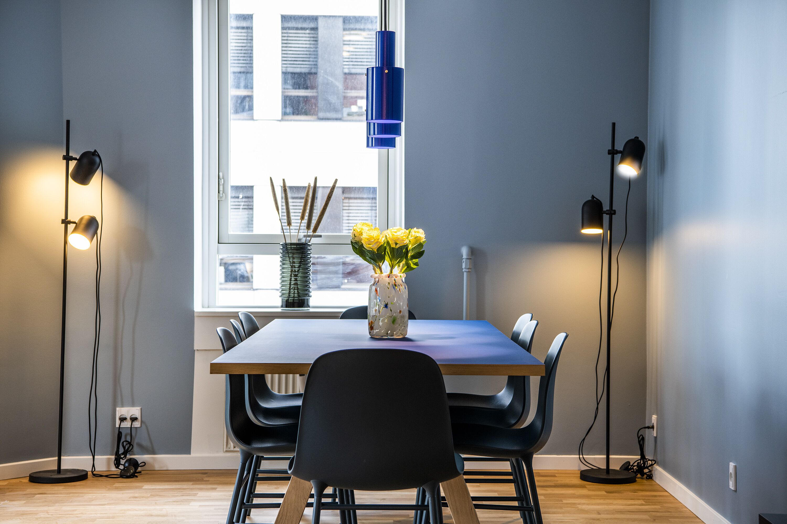 A dining area with a table, chairs, a vase with flowers, and two floor lamps.