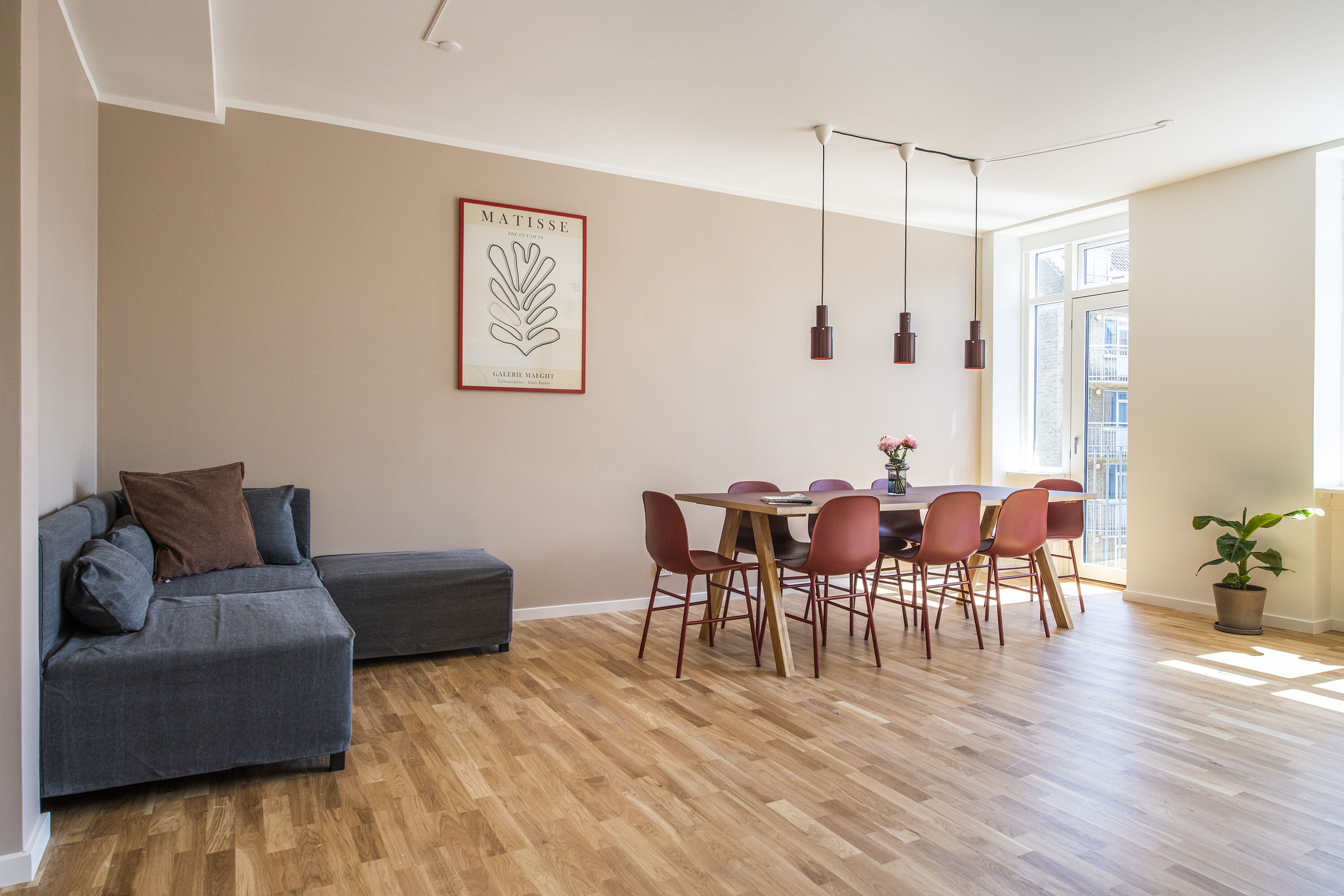 A modern living room with a grey sofa, a dining table with red chairs, wooden flooring, and a large window providing natural light.