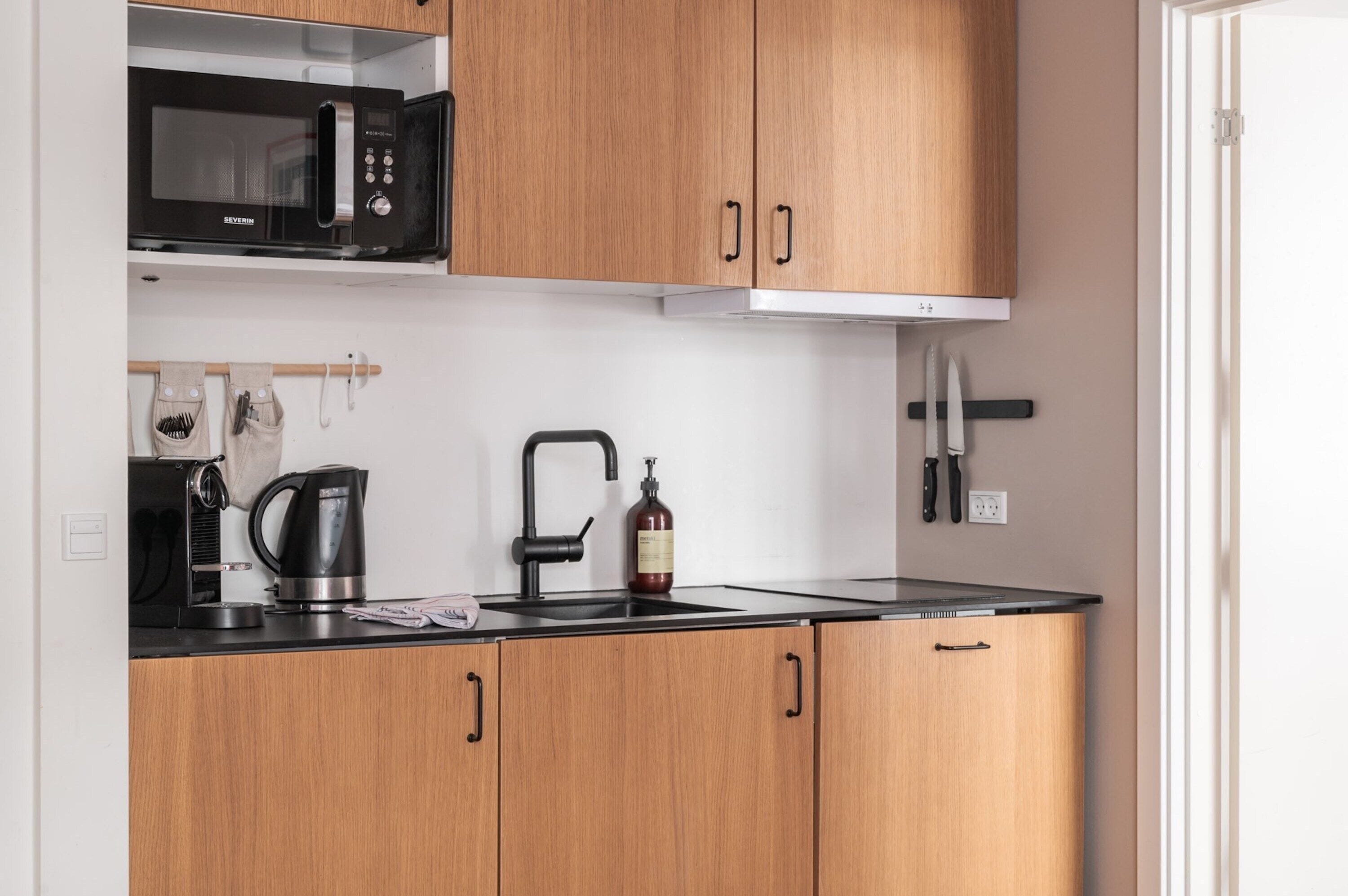 A compact kitchen with wooden cabinets, a black microwave, a kettle, and a sink.
