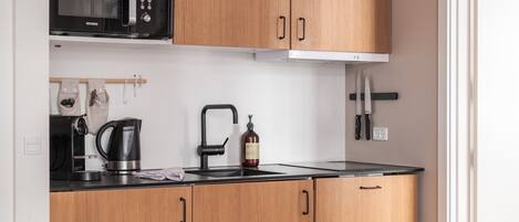 A compact kitchen with wooden cabinets, a black microwave, a kettle, and a sink.