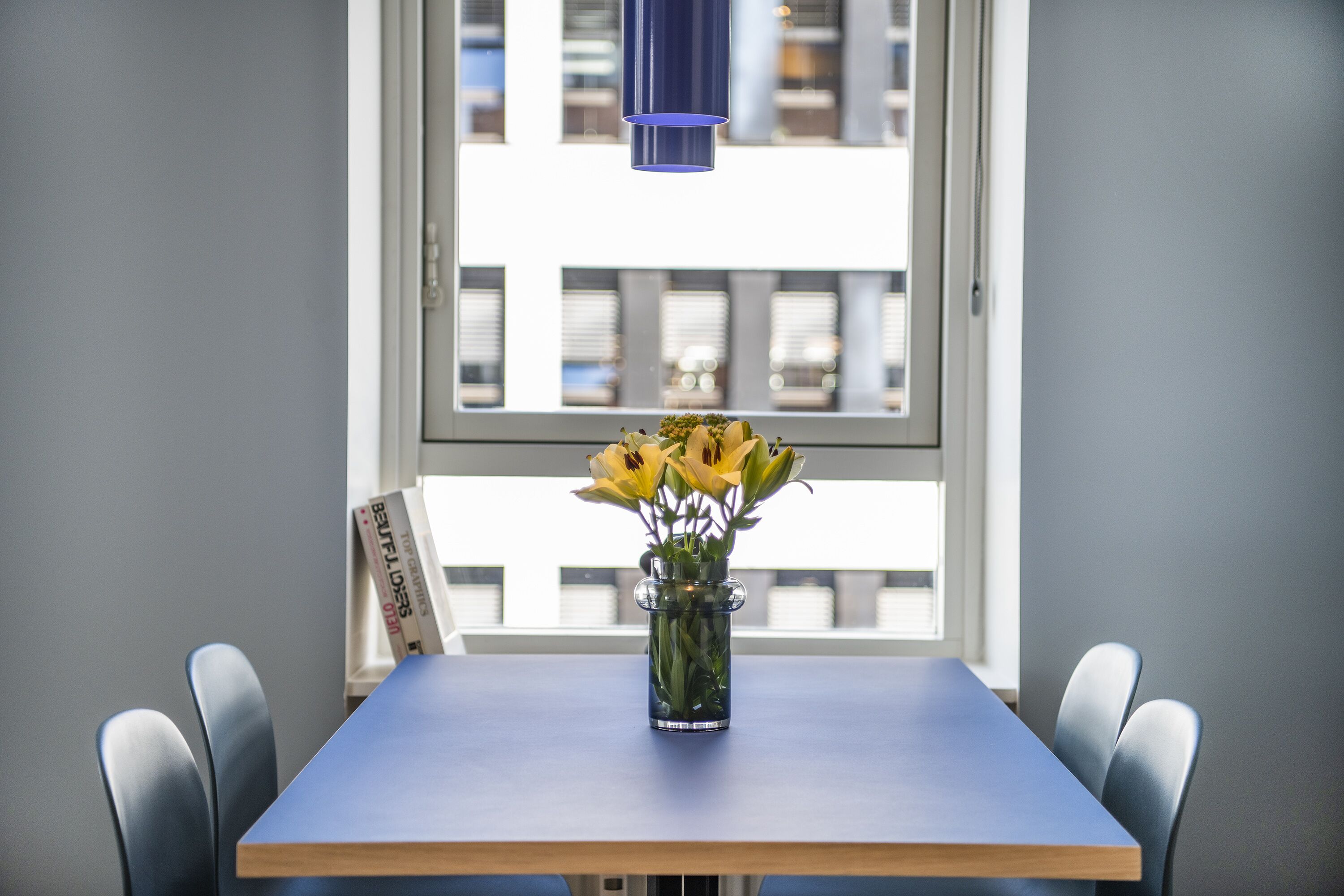 A table with a vase of yellow flowers, a blue tablecloth, and a window with blinds in the background.