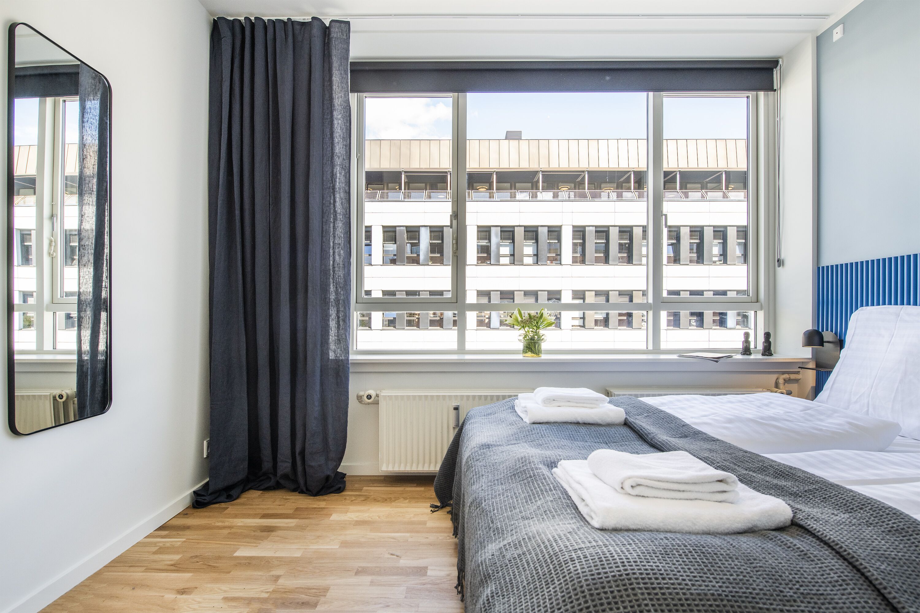 Et hotelværelse med et stort vindue, en seng med pænt foldede håndklæder, en radiator og en potteplante på vindueskarmen.