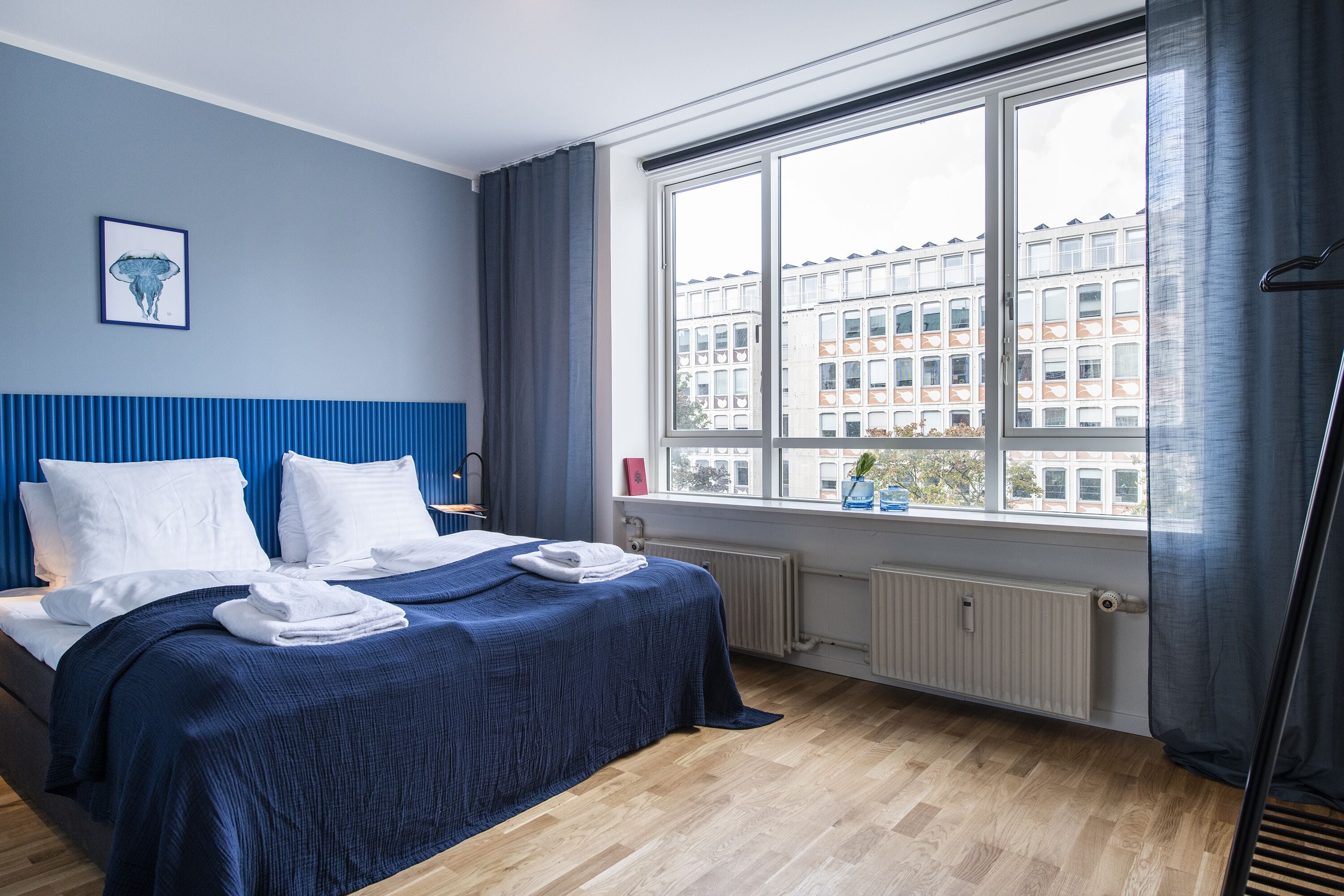 A hotel room with a large window, a bed with white linens, a blue headboard, and a radiator.
