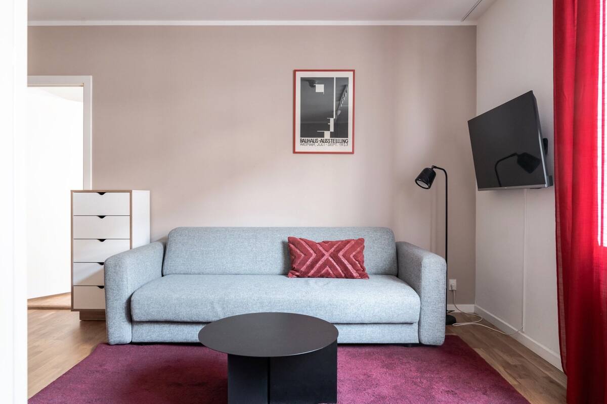 Un salon moderne avec un canapé gris, un coussin rouge, une table basse noire, un téléviseur fixé au mur et un tableau encadré au-dessus du canapé.