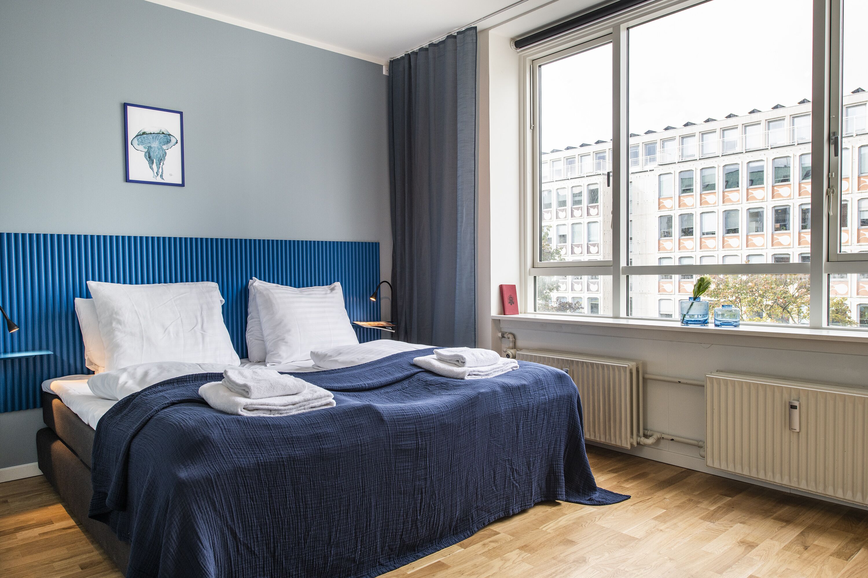 A hotel room with a bed, a window with a view of buildings, a radiator, and a framed picture on the wall.