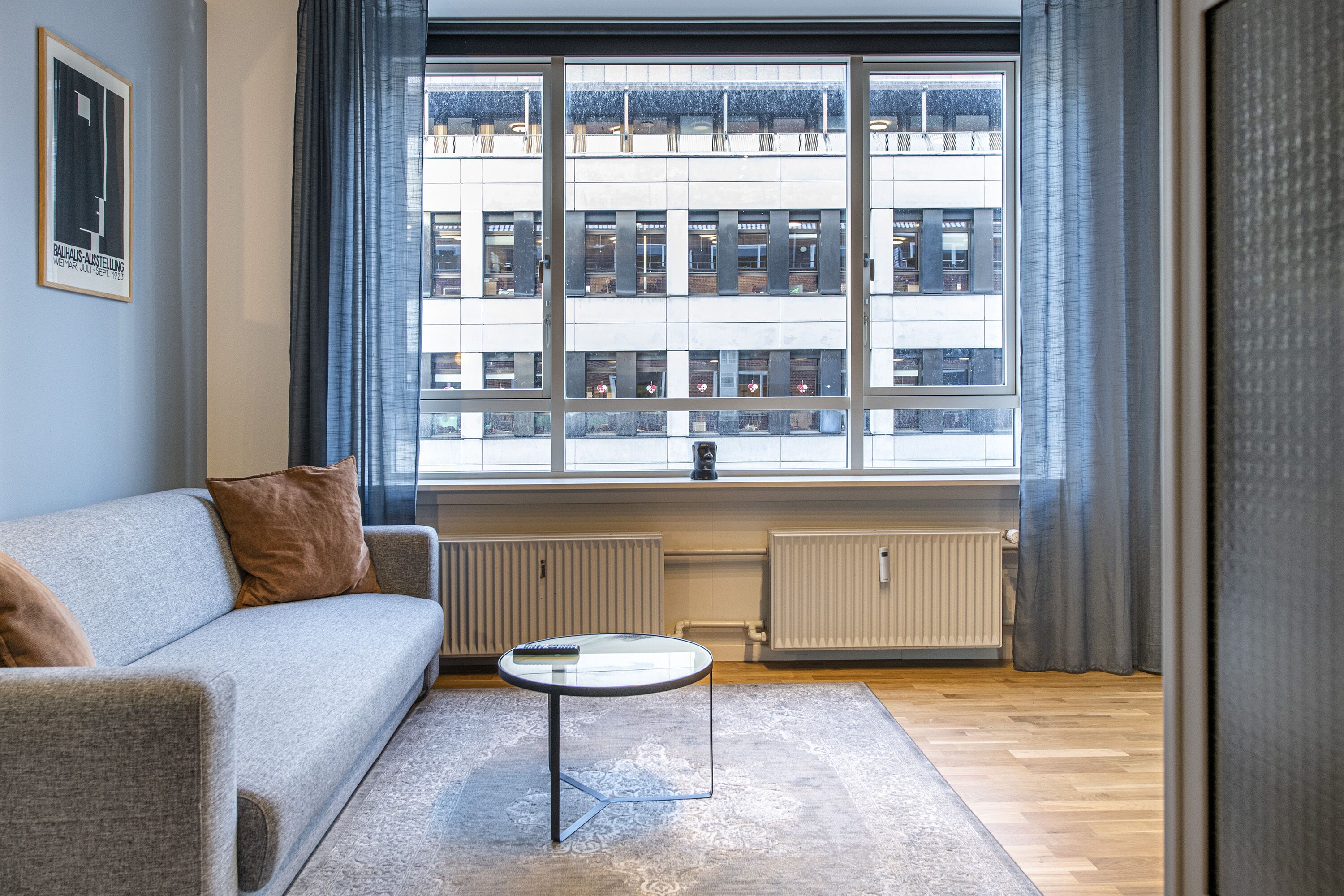 A modern living room with a grey sofa, a glass coffee table, and a view of a building through large windows.