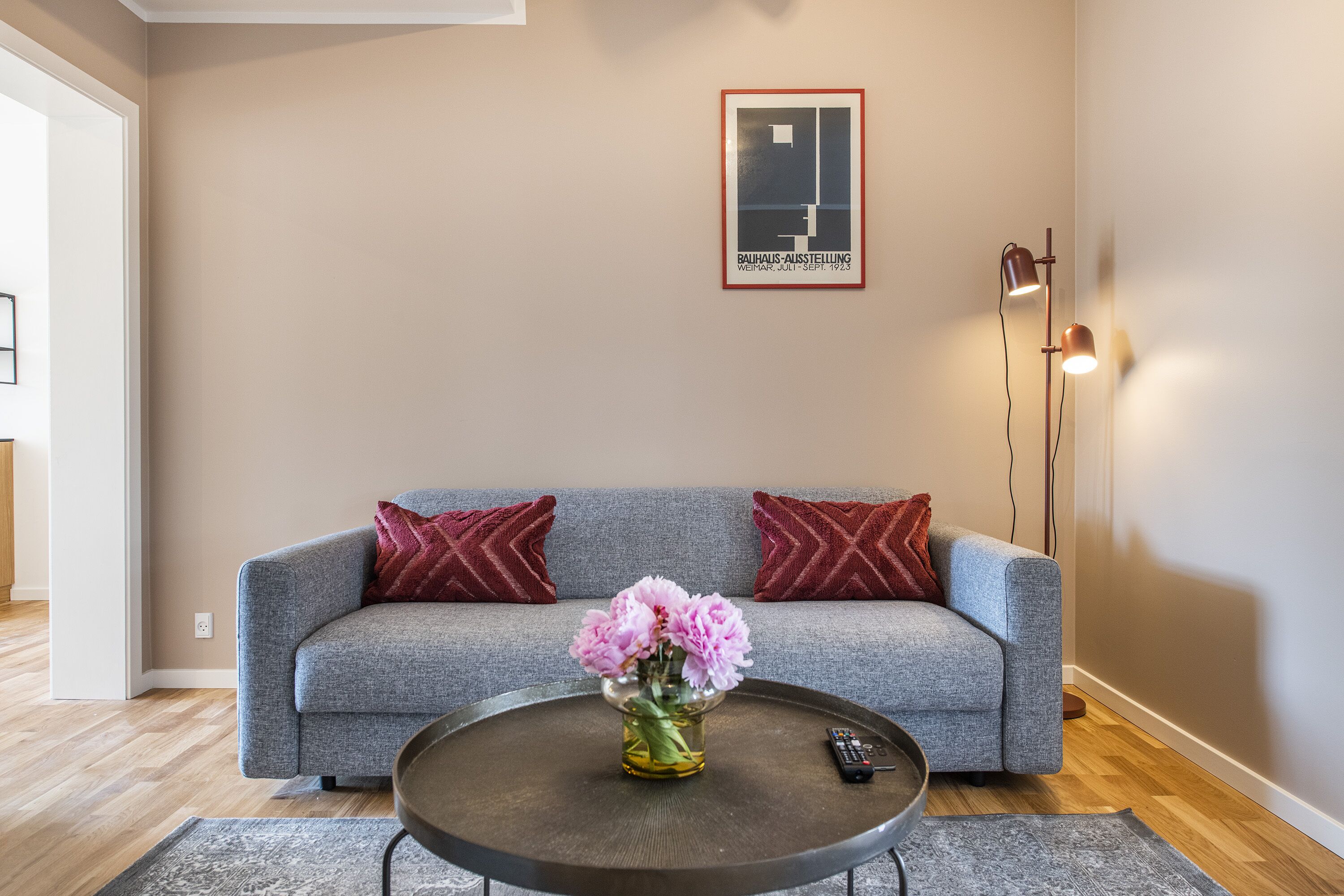 En stue med en grå sofa, to røde puder, en vase med lyserøde blomster på et rundt sofabord, og et indrammet billede på væggen.