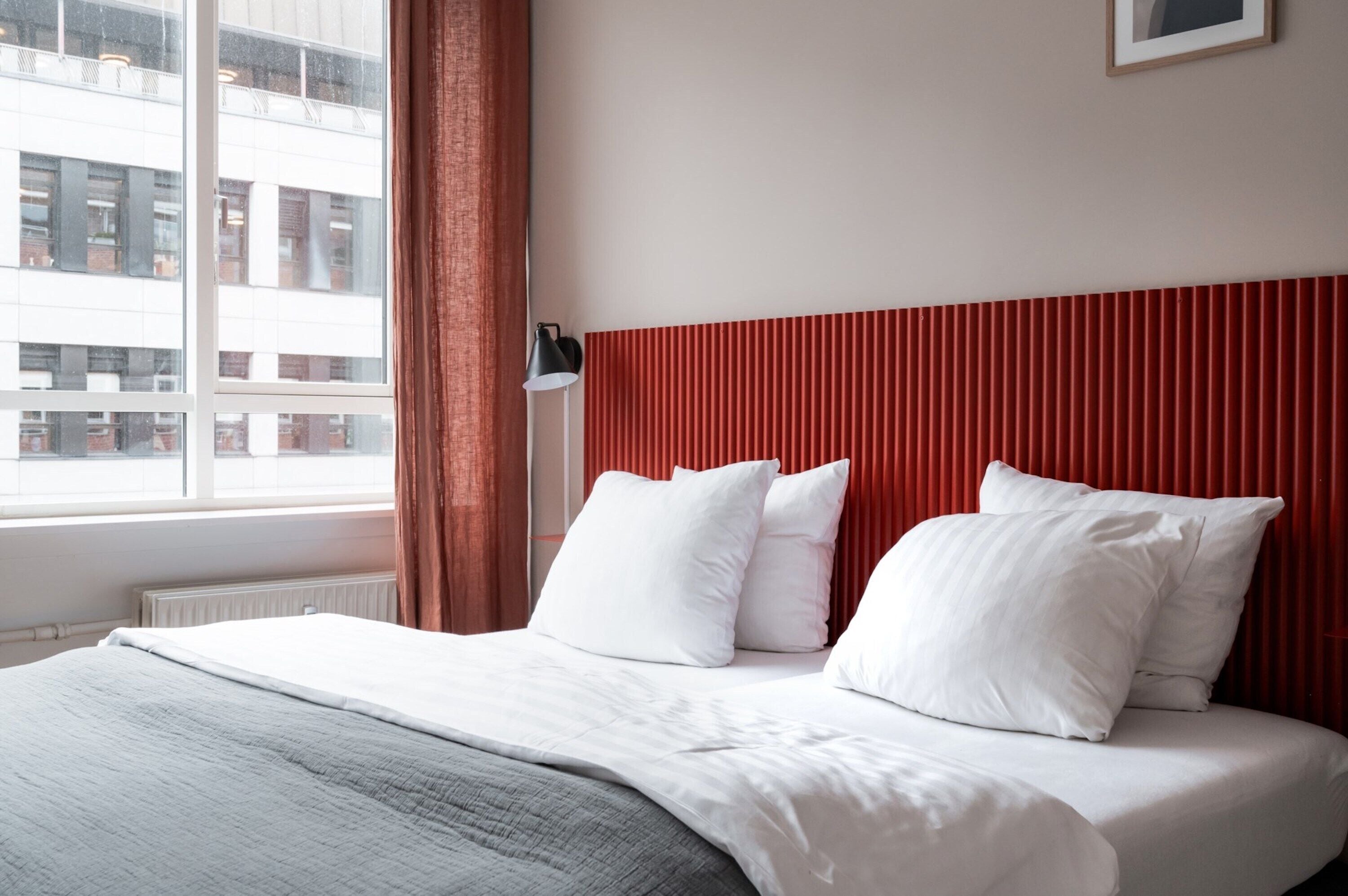 A hotel room with a bed, white bedding, a red headboard, and a window with a view of buildings.