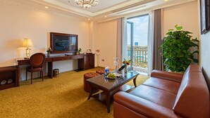 Hotelkamer met een bank, een eettafel, een televisie, een bureau, een stoel, een plant en een balkon met uitzicht.
