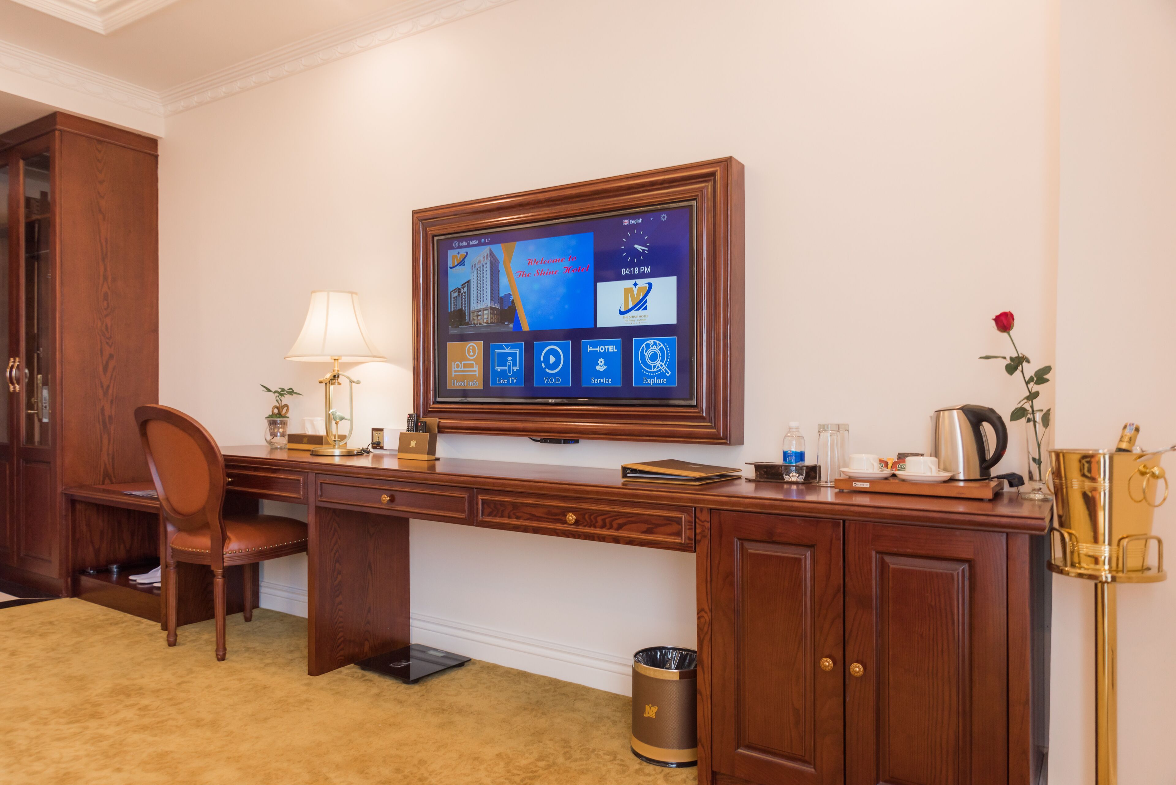 Habitació d'hotel amb escriptori de fusta, un monitor d'ordinador, una làmpada, una cadira, un safataire i una màquina de cafè.