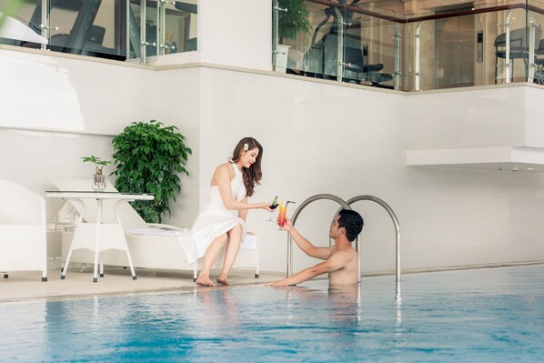 A man and a woman by an indoor pool with a glass railing, a white table, and chairs. - The Shine Hotel (Hai Phong)