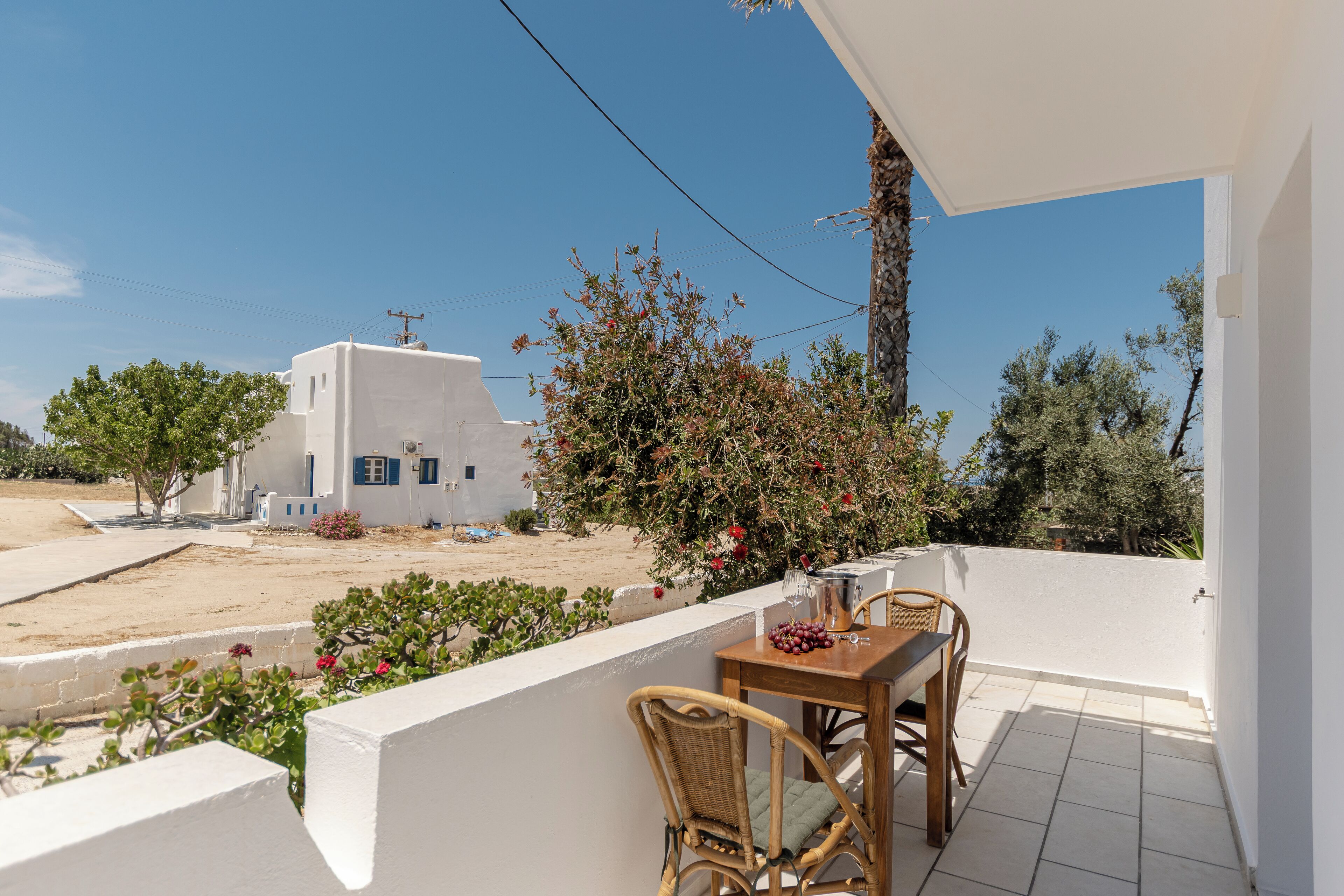 A balcony with a table and chairs, overlooking a white building and a sandy area with trees.