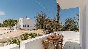 A balcony with a table and chairs, overlooking a white building and a sandy area with trees.