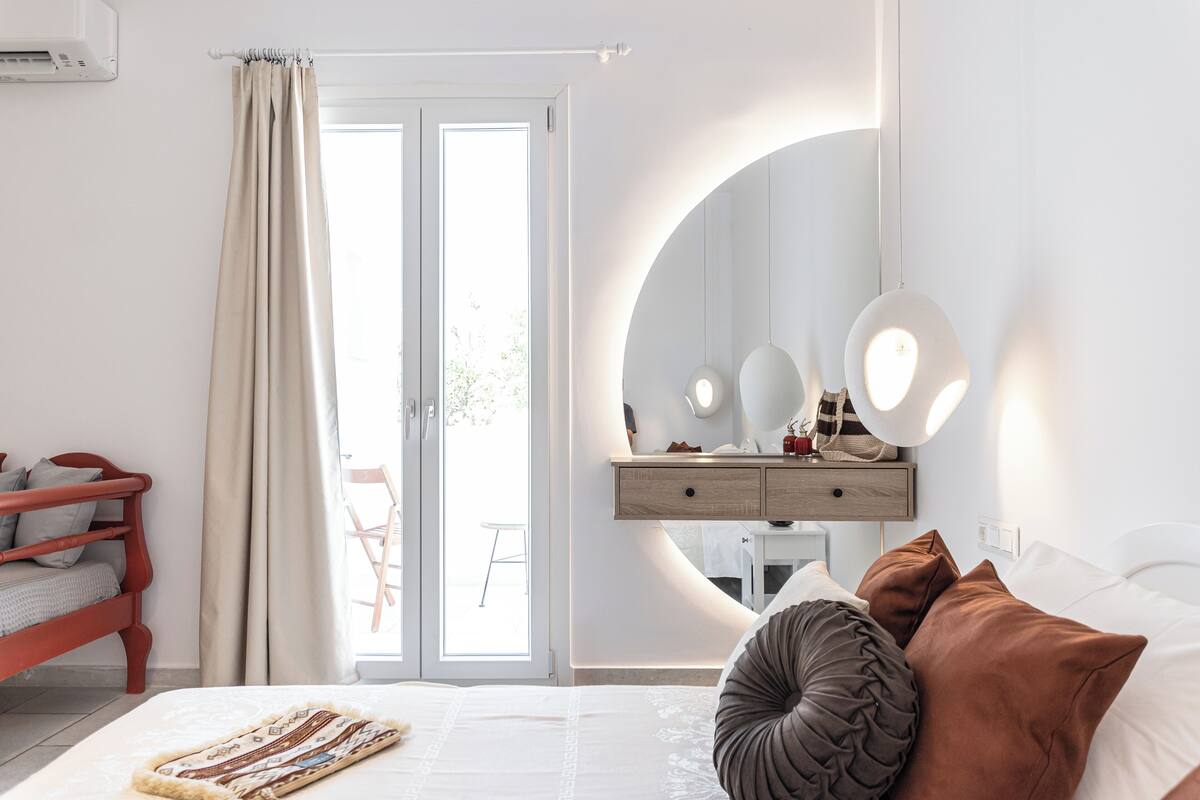 A bedroom with a bed, a bedside table with a mirror, a dresser, and a window with curtains.