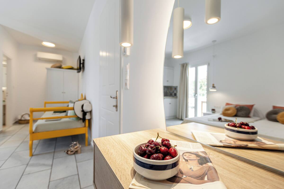 A modern bedroom with a wooden bed, a bedside table with a bowl of cherries, and a large mirror reflecting the room.