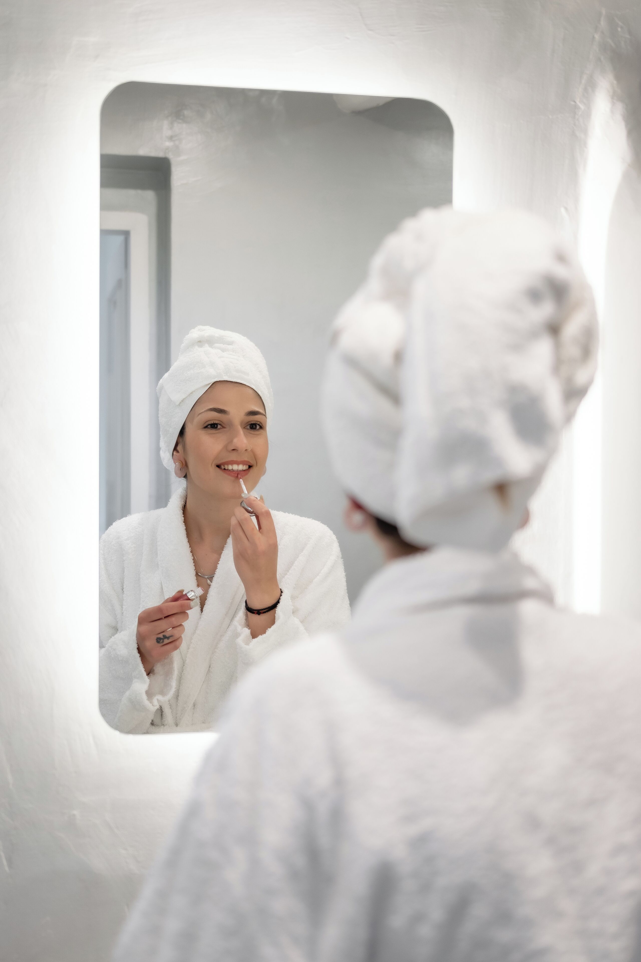 A person in a white bathrobe with a towel wrapped around their head, standing in front of a mirror.