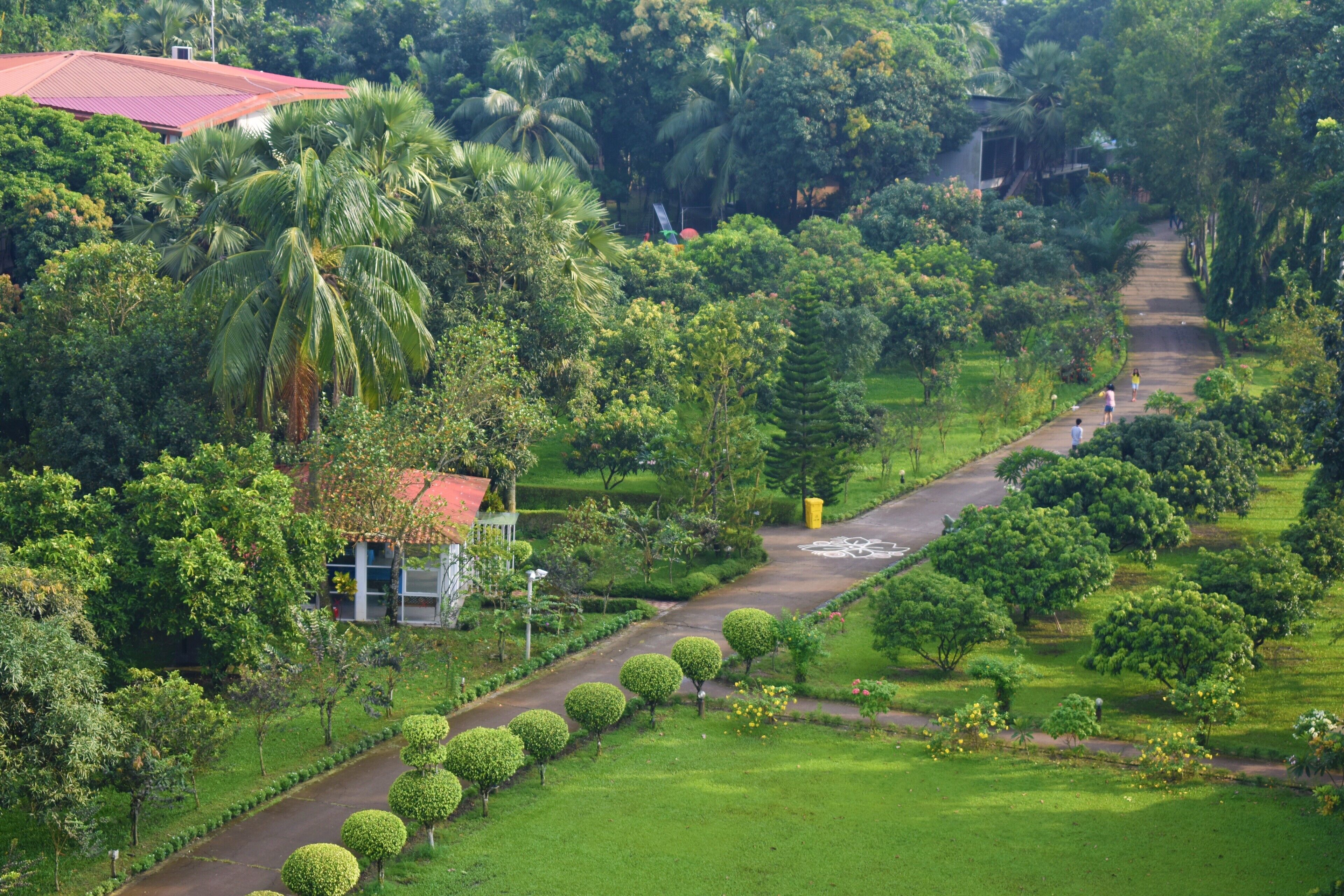 A lush garden with a pathway, a building with a red roof, and a small structure with a glass facade.