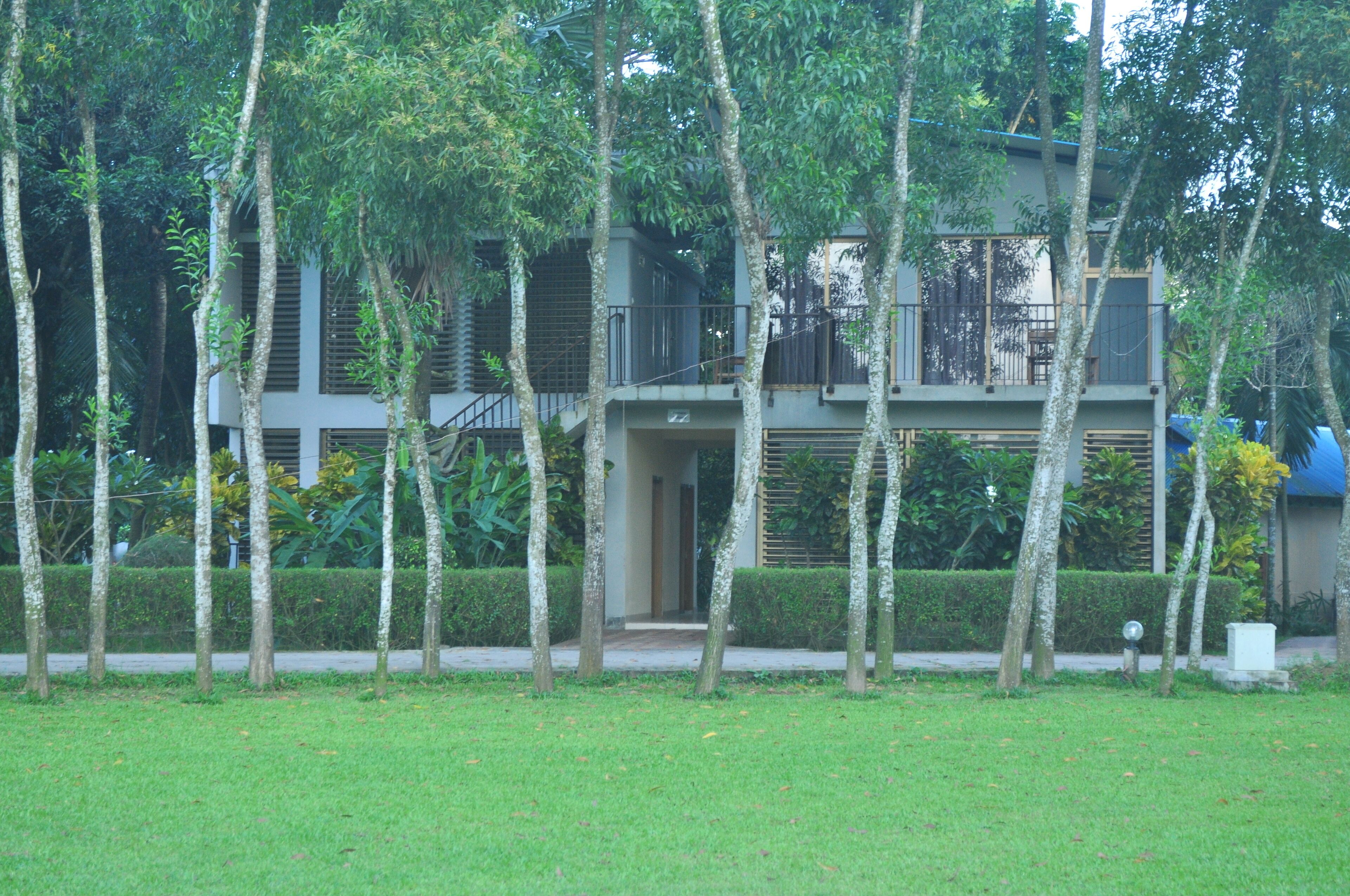 A two-story building with a balcony and a covered walkway, surrounded by trees and a grassy area.