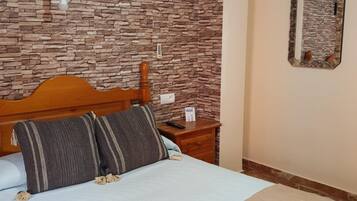 Une chambre avec un mur décoré d’un motif imitant la pierre, une tête de lit en bois, un lit agrémenté d’oreillers à rayures, une table de chevet avec une télécommande et un miroir sur le mur.