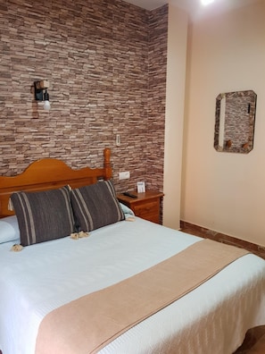 Un dormitorio con pared de diseño de piedra, cabecera de madera, cama con almohadas a rayas, mesita de noche con control remoto y espejo en la pared.