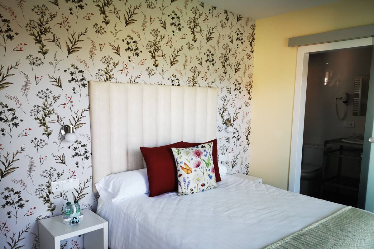 A neatly made bed with white bedding and two pillows, a small bedside table with a lamp, and a floral wallpaper pattern.