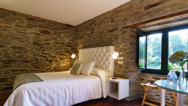 A bedroom with a stone wall, a bed with white bedding, a bedside table, and a small round table with a vase.