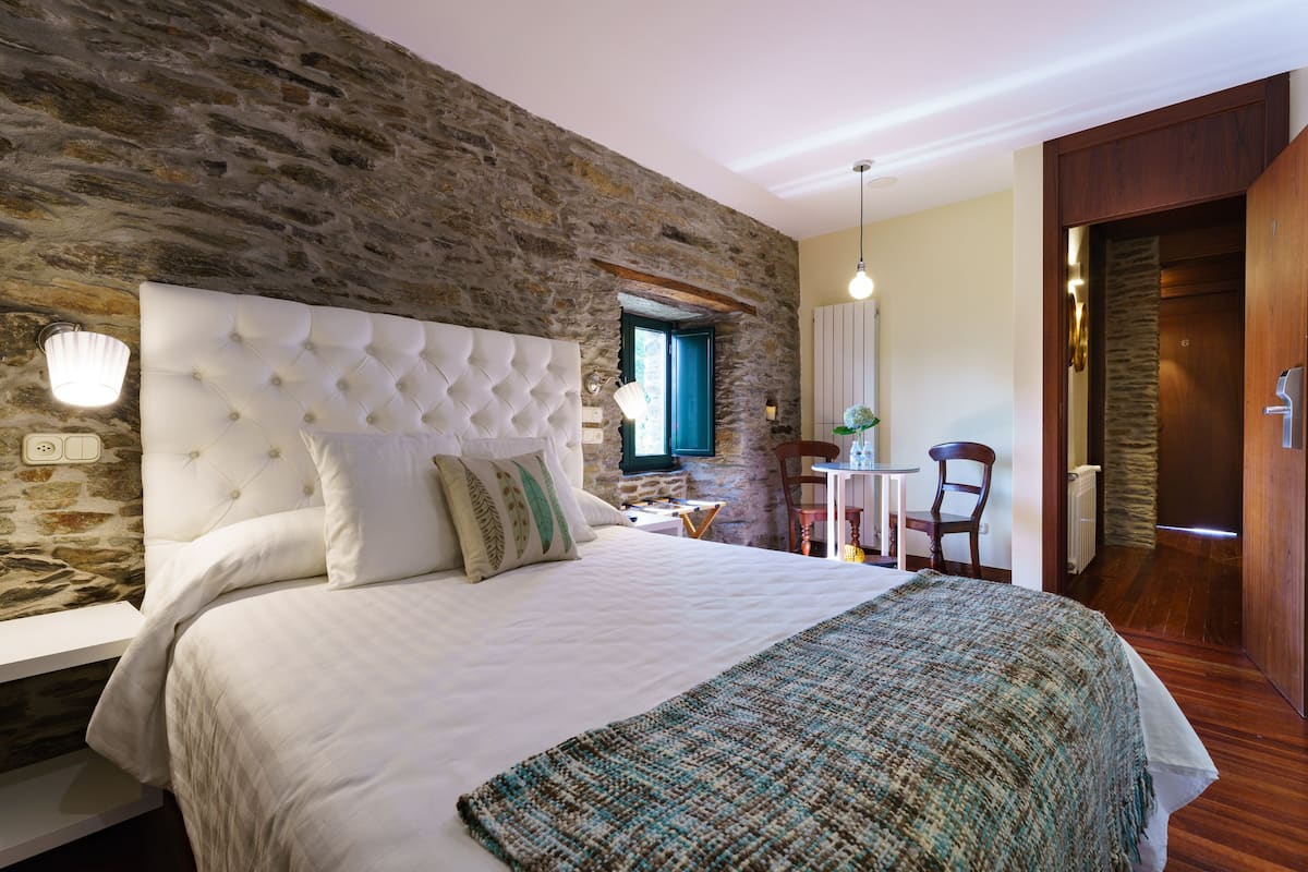 A bedroom with a stone wall, a tufted headboard, a white bedspread, and a woven throw blanket.
