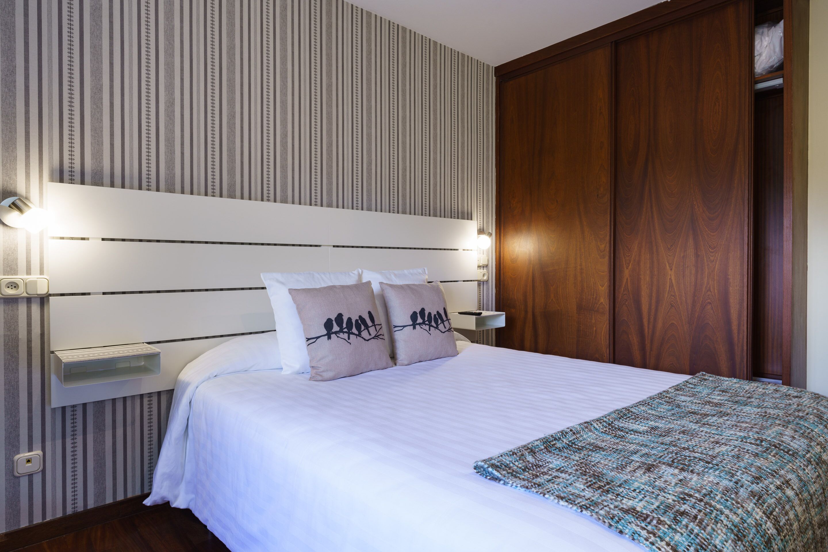 Een net opgemaakt bed met een witte dekbedovertrek en twee kussens met vogelmotieven, een nachtkastje met een lamp en een houten garderobe.