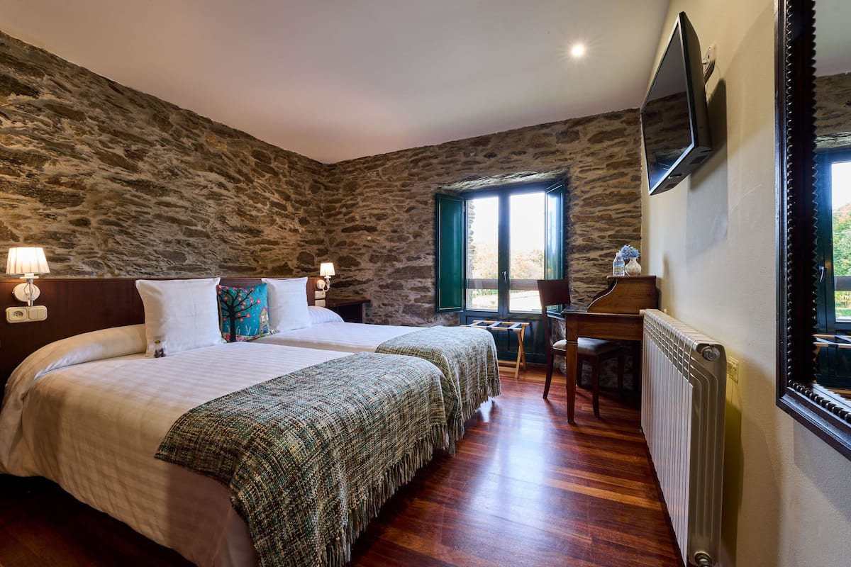 A bedroom with a stone wall, a bed with white bedding, a green blanket, a wooden chair, a small table, and a TV mounted on the wall.