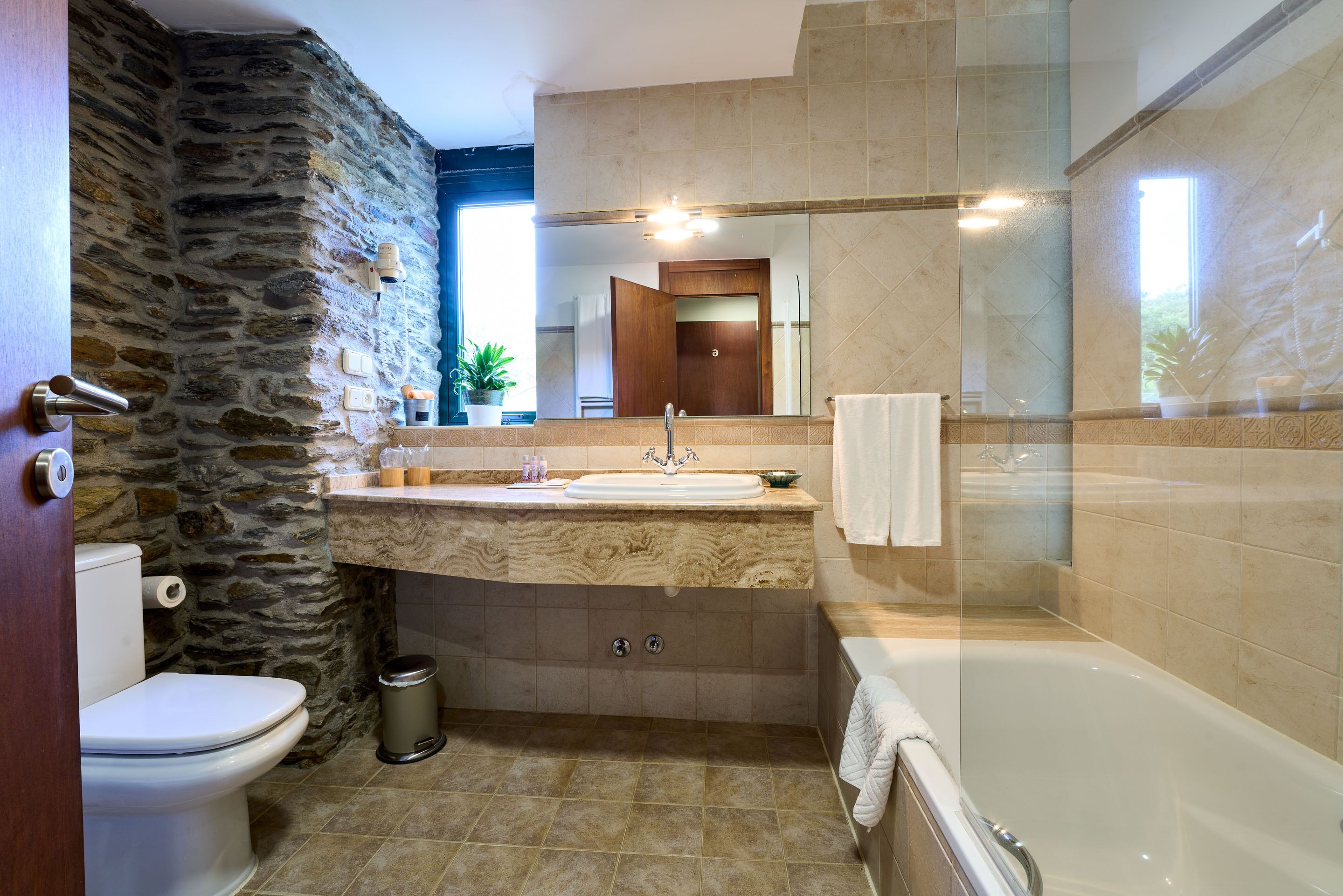 A bathroom with a stone wall, a marble sink, a toilet, a trash can, and a bathtub.