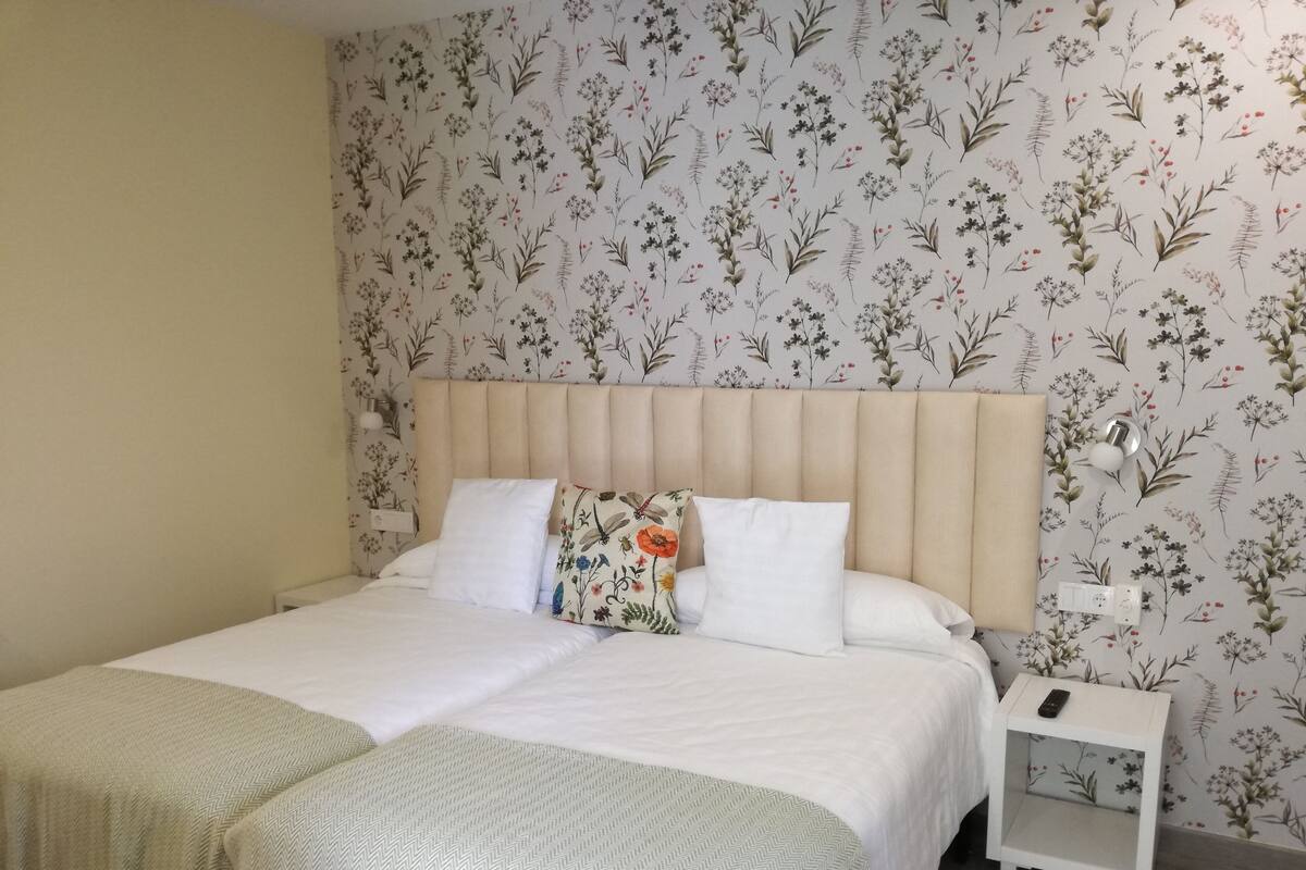 A neatly made bed with a white headboard, a bedside table with a remote control, and a wall with floral wallpaper.