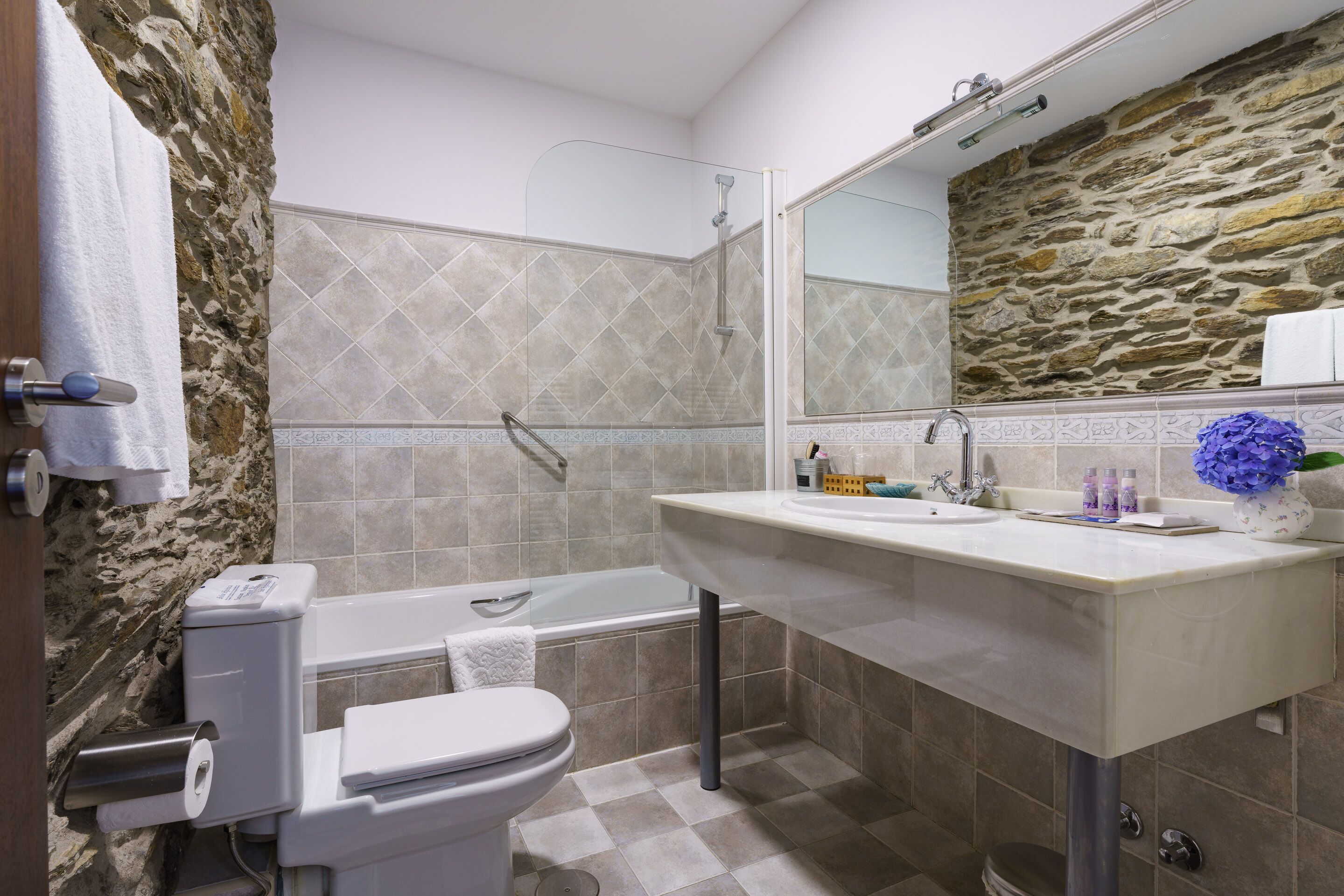 Een badkamer met een stenen muur, een bad, een toilet en een wastafel met spiegel.