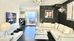 Un salon moderne avec un canapé blanc, une table basse et un téléviseur fixé au mur. Il y a un balcon avec vue sur la mer.