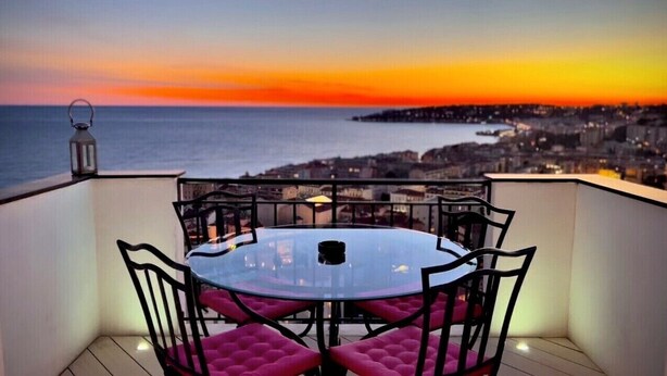 Une table ronde en verre, entourée de chaises en métal noir et agrémentée de coussins roses, sur un balcon donnant sur une vue panoramique de la ville au coucher du soleil.